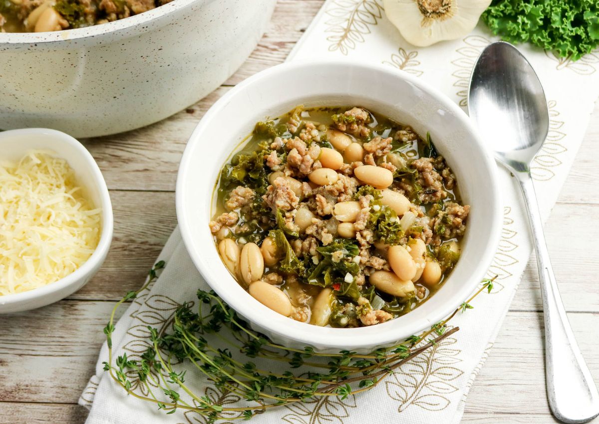 A white bowl of soup with white beans, ground meat, kale, and broth, placed on a napkin with a spoon beside it; shredded cheese and fresh herbs are nearby.