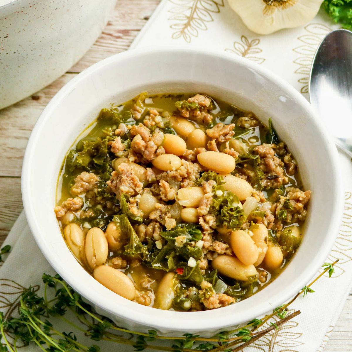 A white bowl filled with white bean and sausage soup with kale, placed on a patterned napkin next to a spoon and fresh herbs.