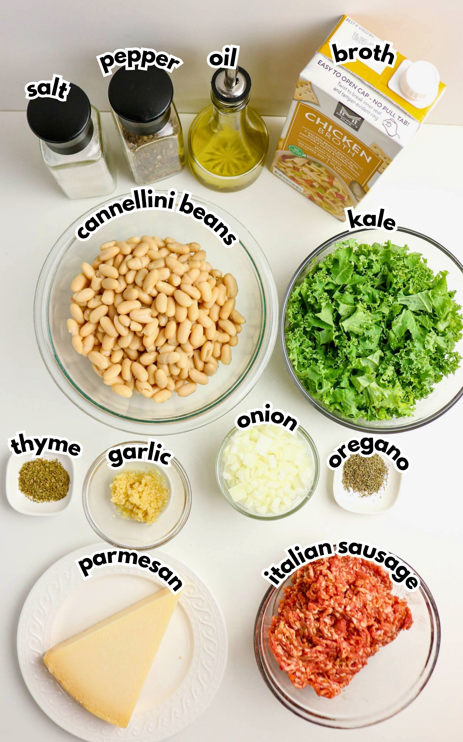Ingredients for a soup recipe are arranged on a white surface, including cannellini beans, kale, italian sausage, broth, onion, garlic, spices, oil, salt, pepper, and parmesan cheese.