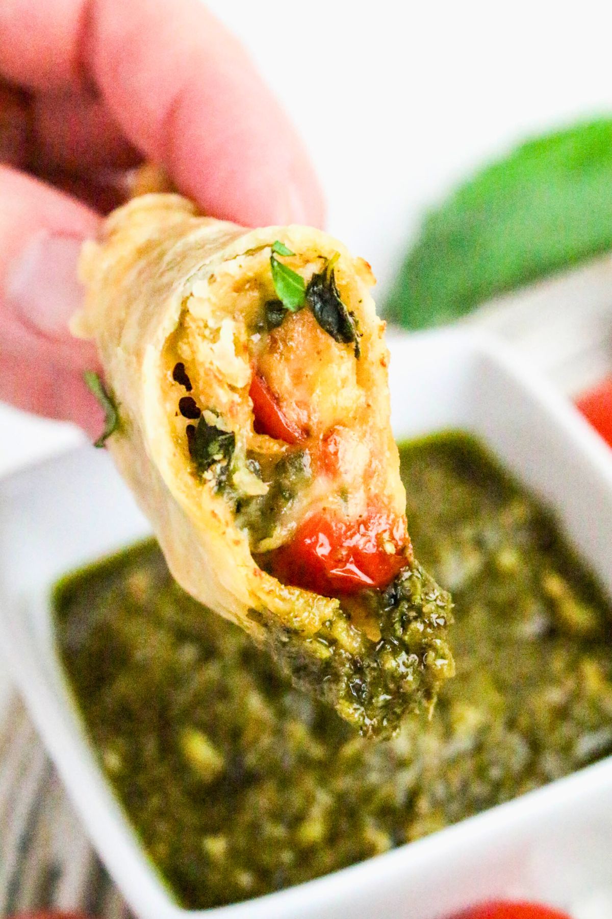 A hand holds a pastry filled with tomatoes, spinach, and cheese, with a bowl of green dipping sauce in the background.