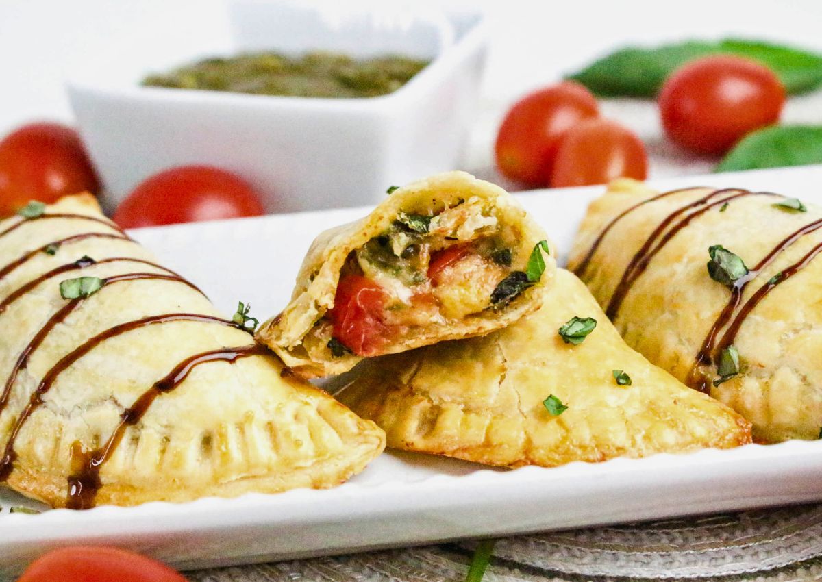 Three baked empanadas with golden crust on a plate, one cut open to show a vegetable filling, garnished with chopped herbs and balsamic glaze; blurred tomatoes and sauce in the background.