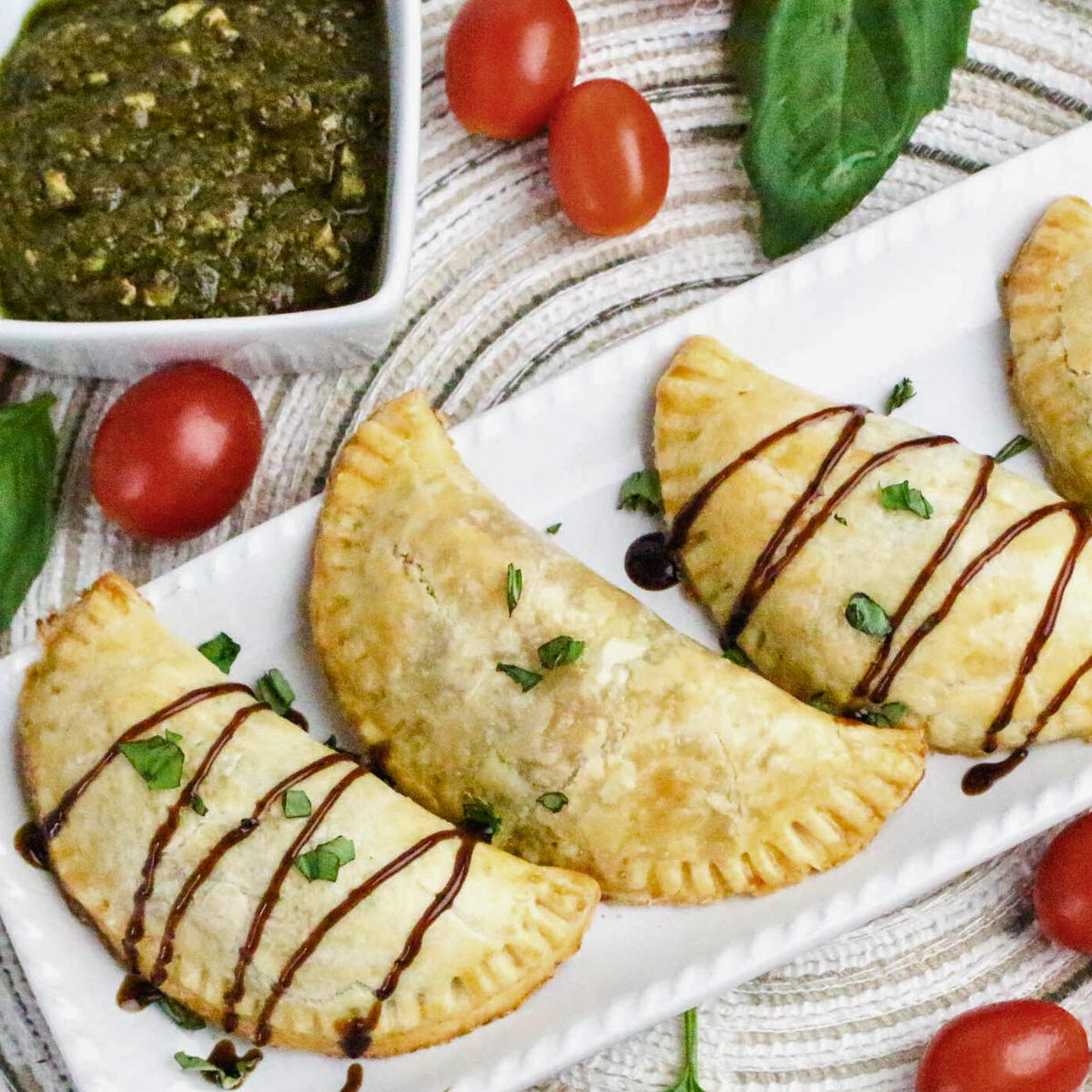 Three baked empanadas drizzled with sauce and garnished with herbs are served on a white platter, with cherry tomatoes, basil leaves, and a bowl of pesto dip nearby.