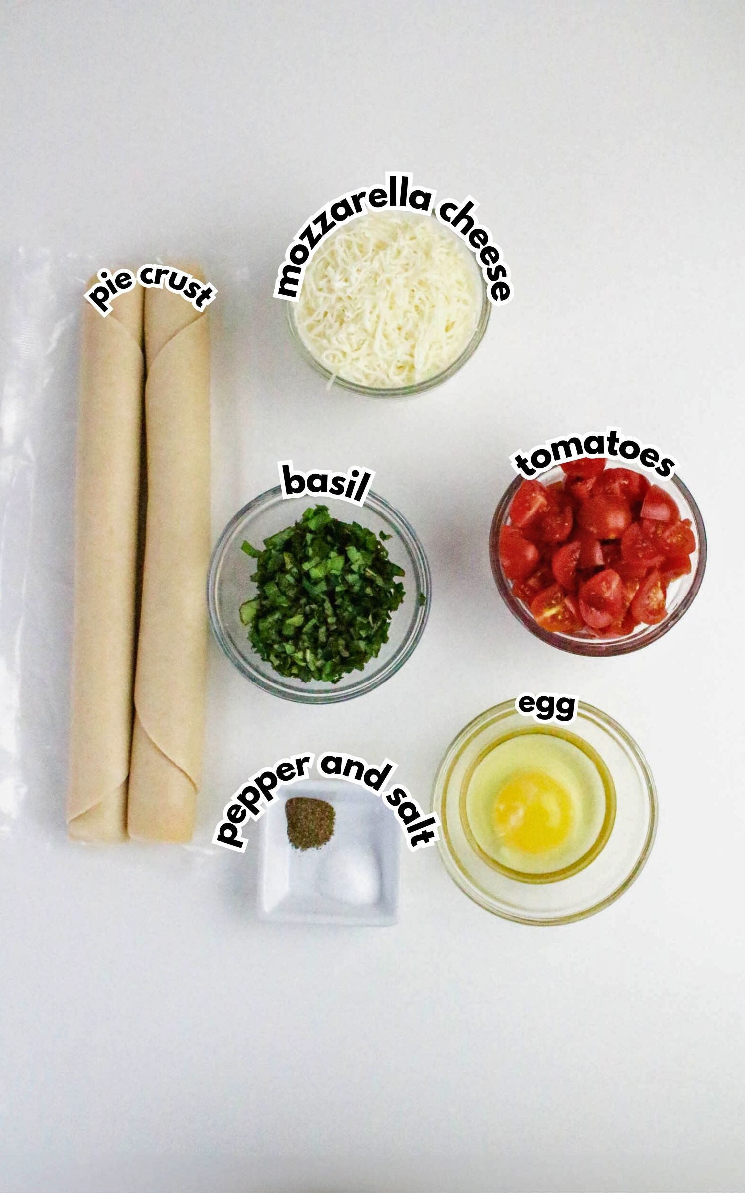 Two rolled pie crusts, shredded mozzarella cheese, chopped tomatoes, chopped basil, a dish with pepper and salt, and a bowl with a cracked egg are arranged on a white surface.