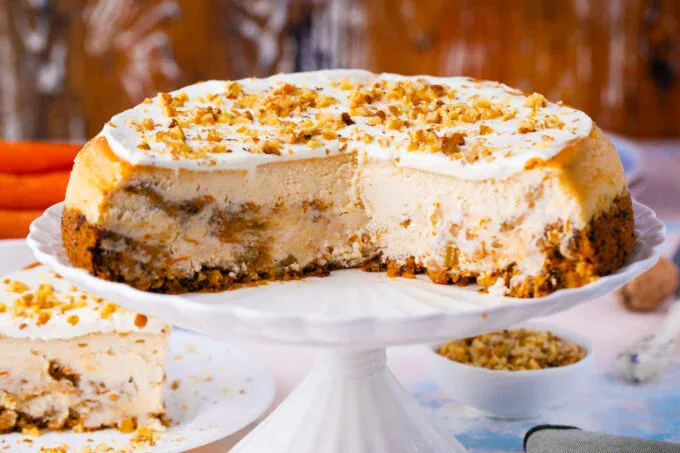 A carrot cake cheesecake with a slice removed, showing creamy layers and a crumbly base, topped with frosting and chopped nuts on a white cake stand.