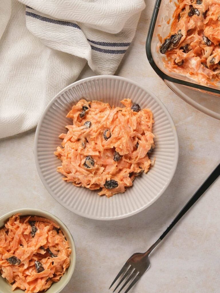 A bowl of carrot salad with raisins and creamy dressing sits next to a fork, a smaller bowl, and a glass dish of the same salad on a light surface.