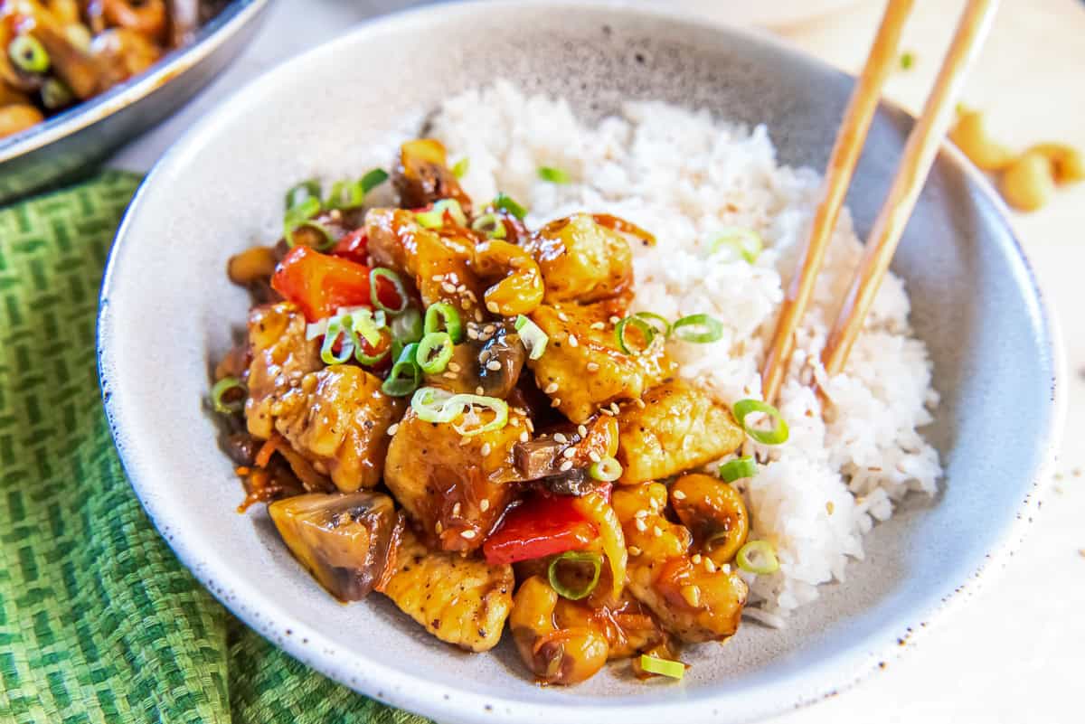A bowl of white rice topped with stir-fried chicken, mushrooms, red bell peppers, and green onions, garnished with sesame seeds. Chopsticks are resting on the bowl.