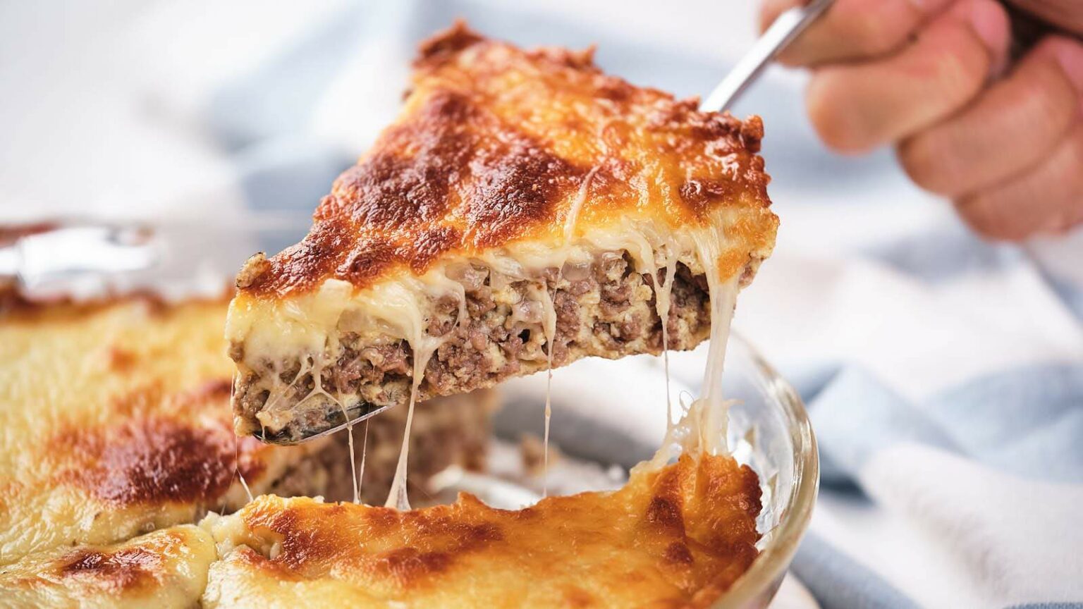 A slice of baked casserole with melted cheese and ground meat is being lifted from a glass dish.