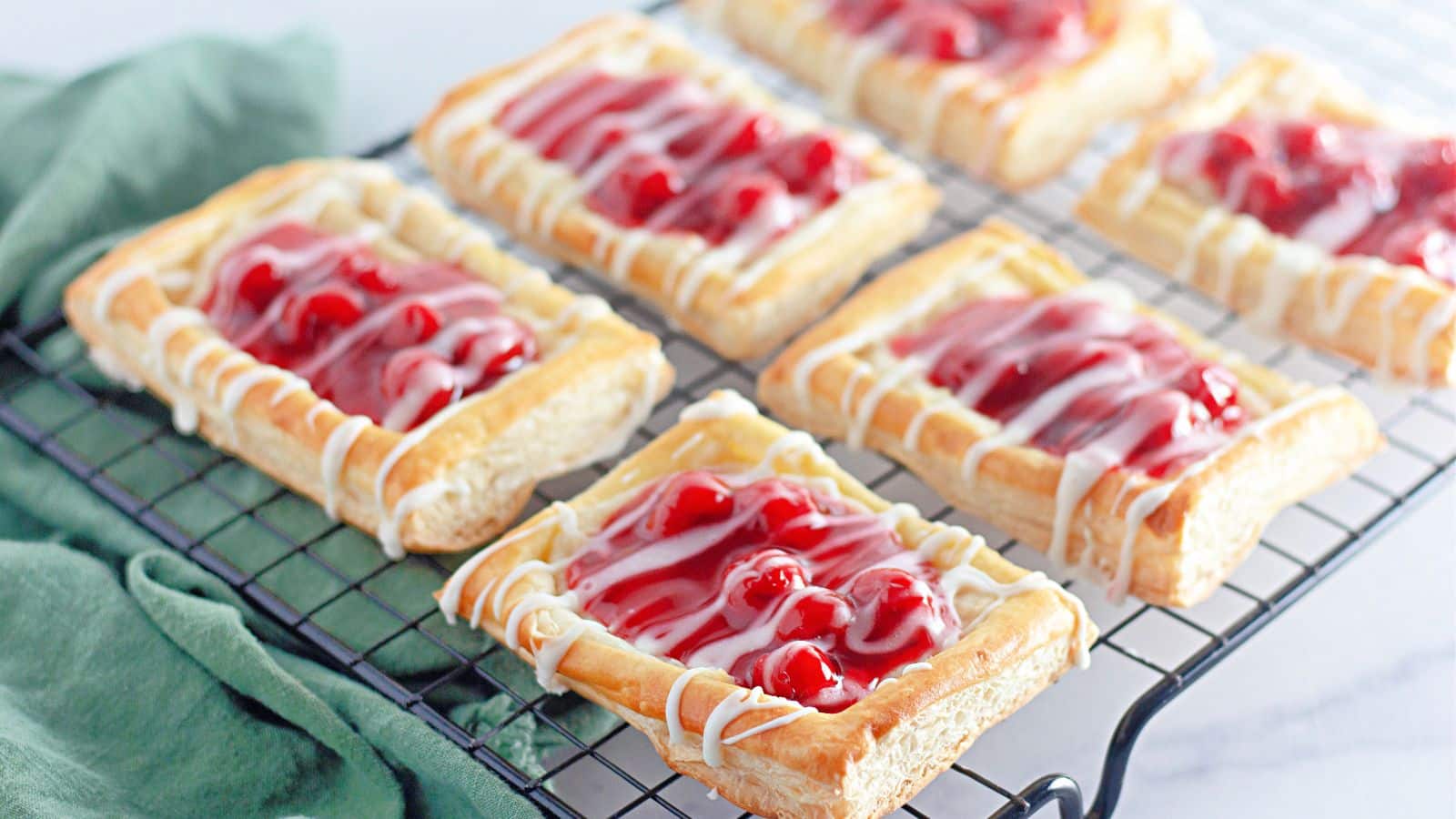 Rectangular cherry danishes with white icing drizzle are cooling on a black wire rack, with a green cloth placed beside them.
