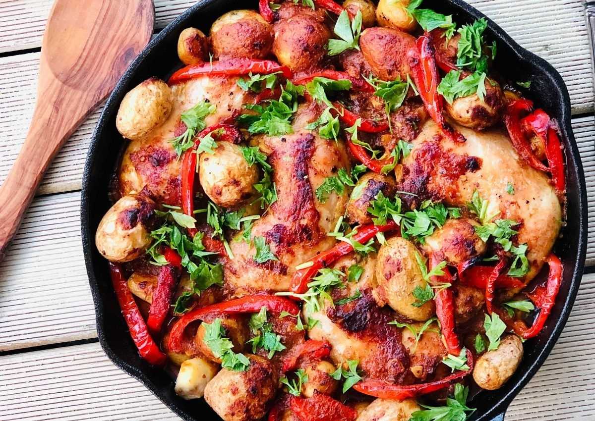 A skillet filled with roasted chicken, potatoes, red bell peppers, and fresh parsley, placed on a wooden surface with a wooden spoon beside it.