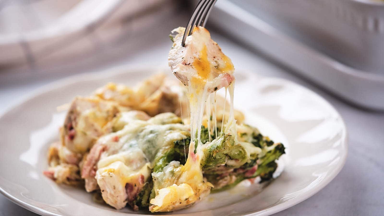 A fork lifts a cheesy baked dish containing pieces of chicken, broccoli, and melted cheese from a white plate.