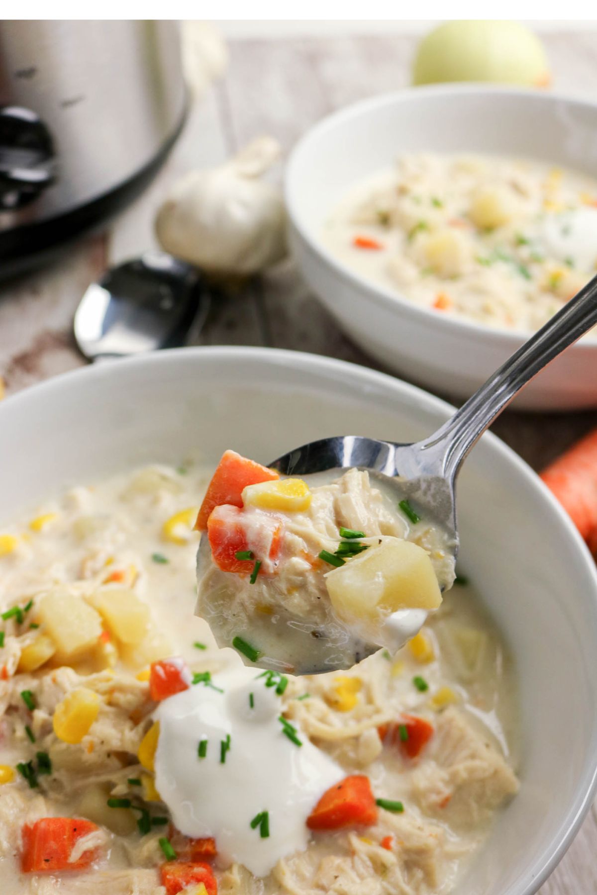 A spoonful of creamy chicken and vegetable soup is held above a bowl, with visible chunks of chicken, potato, corn, and red bell pepper, garnished with sour cream and chives.