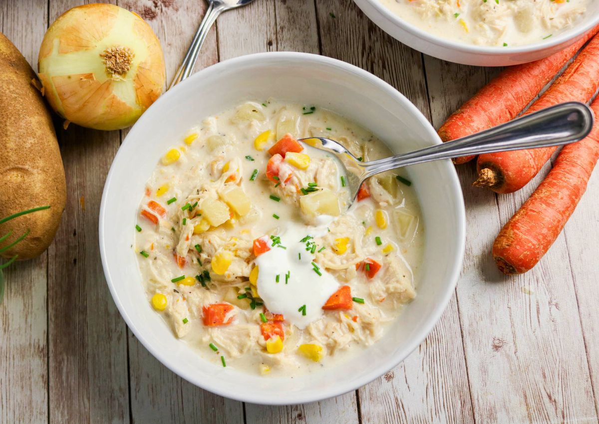 A bowl of creamy chicken and vegetable soup garnished with sour cream and chives, with a spoon inside. Whole carrots, an onion, and a potato are on the wooden surface nearby.