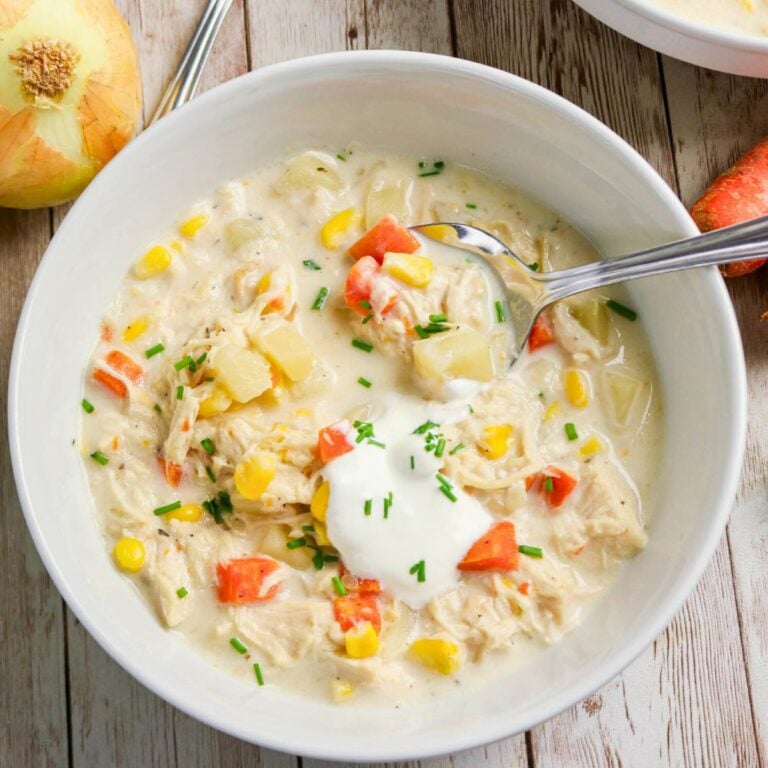 A bowl of creamy chicken and vegetable soup with corn, carrots, potatoes, chives, and a dollop of sour cream, served with a spoon.
