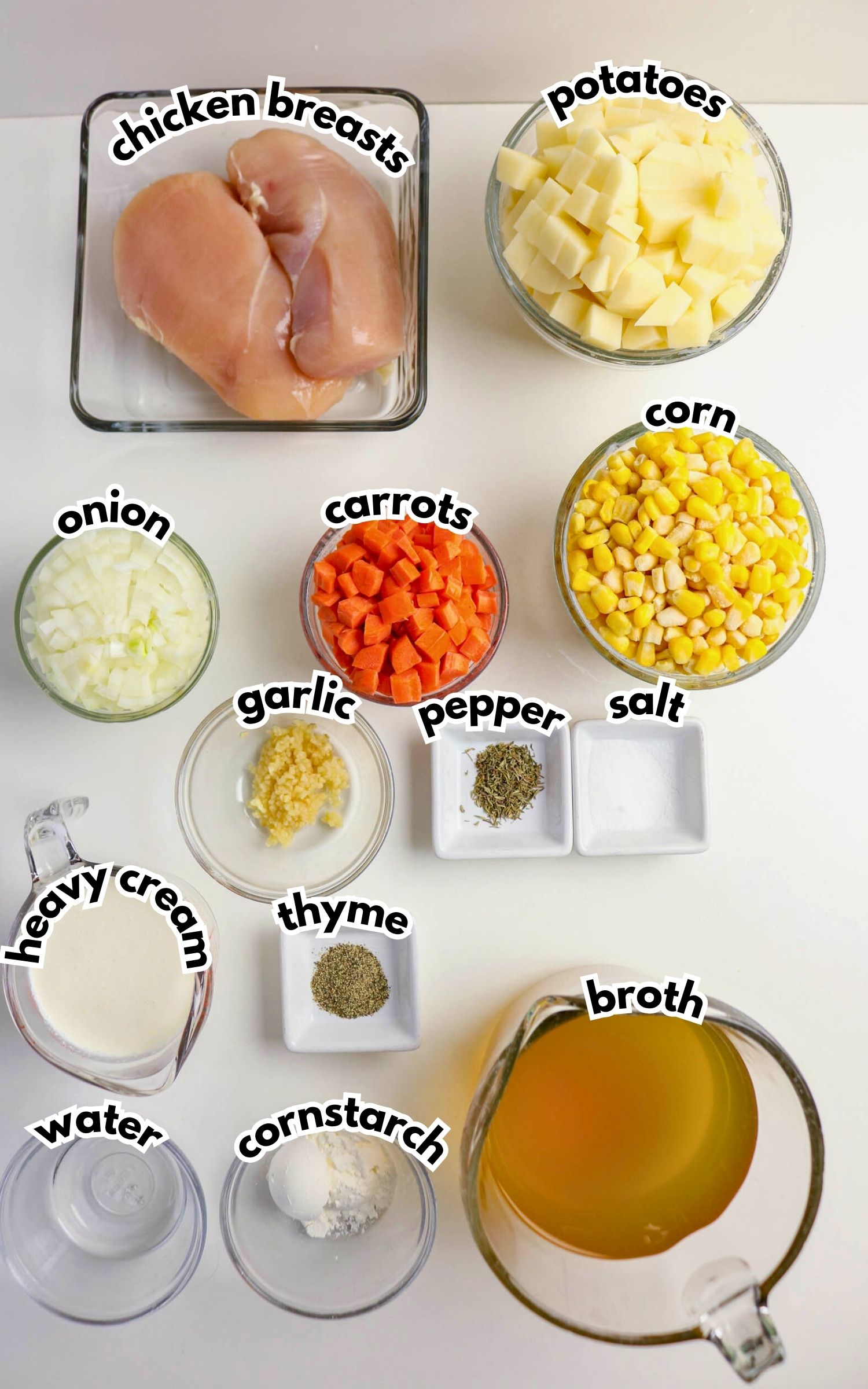 Ingredients for a chicken soup laid out on a white surface, including chicken breasts, potatoes, onions, carrots, corn, cream, water, cornstarch, broth, garlic, pepper, salt, and thyme.