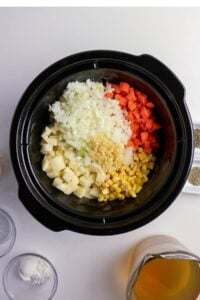 A slow cooker contains chopped onions, carrots, potatoes, corn, and minced garlic, with seasonings and broth nearby.