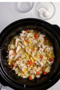 A slow cooker filled with a mixture of shredded chicken, diced potatoes, corn, carrots, and broth. A small bowl of cornstarch and an egg are beside the cooker.