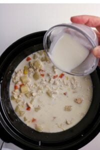 A hand pours a mixture from a small bowl into a slow cooker filled with a creamy soup containing diced vegetables and chicken.