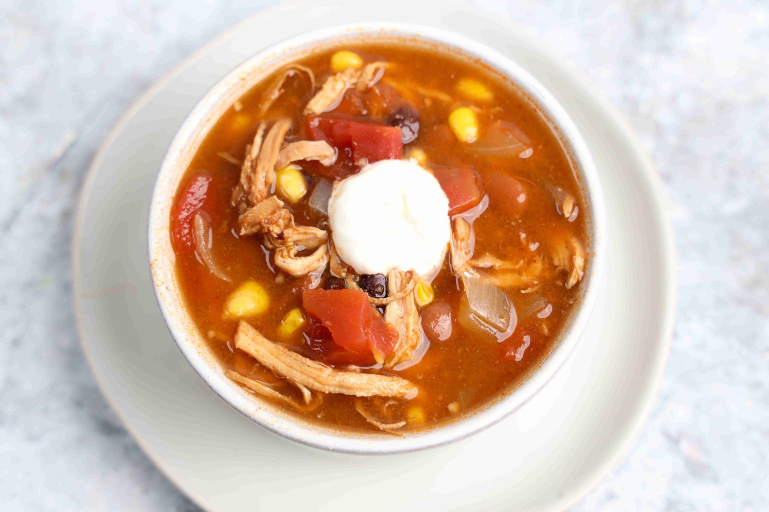 A bowl of chicken tortilla soup with shredded chicken, corn, beans, tomatoes, and a dollop of sour cream on top, served on a white plate.