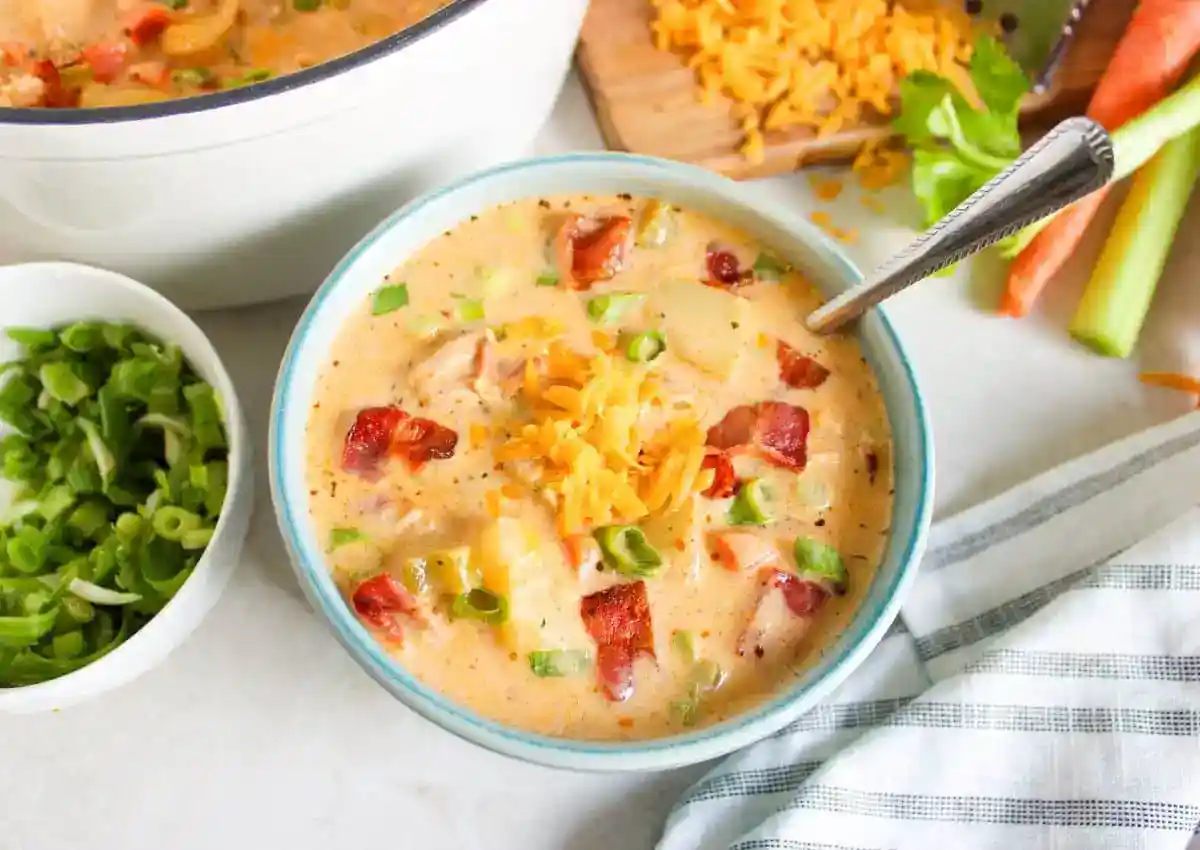A bowl of creamy soup topped with shredded cheese, bacon bits, and chopped green onions, with a spoon inside. Nearby are a bowl of green onions and a cutting board with cheese and vegetables.