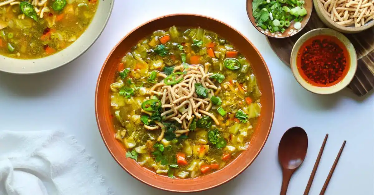A bowl of vegetable soup garnished with crunchy noodles and herbs, surrounded by bowls of chopped green onions, red chili flakes, and utensils on a white surface.
