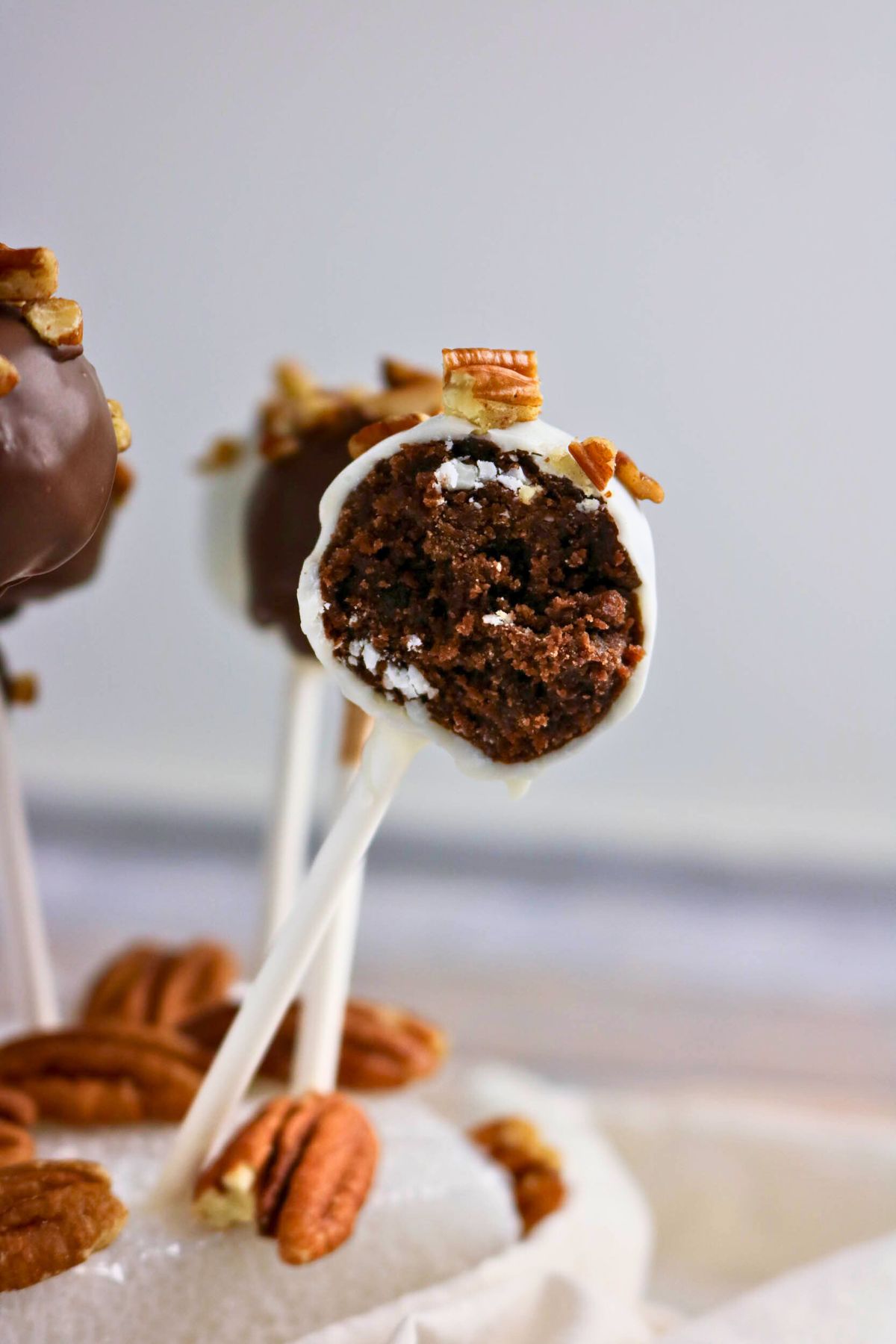 A close-up of a chocolate cake pop with a bite taken out, coated in white chocolate and topped with chopped pecans, on a white background.