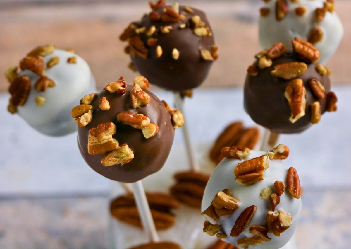 Six cake pops coated in white and milk chocolate, topped with chopped pecans, displayed upright on sticks.