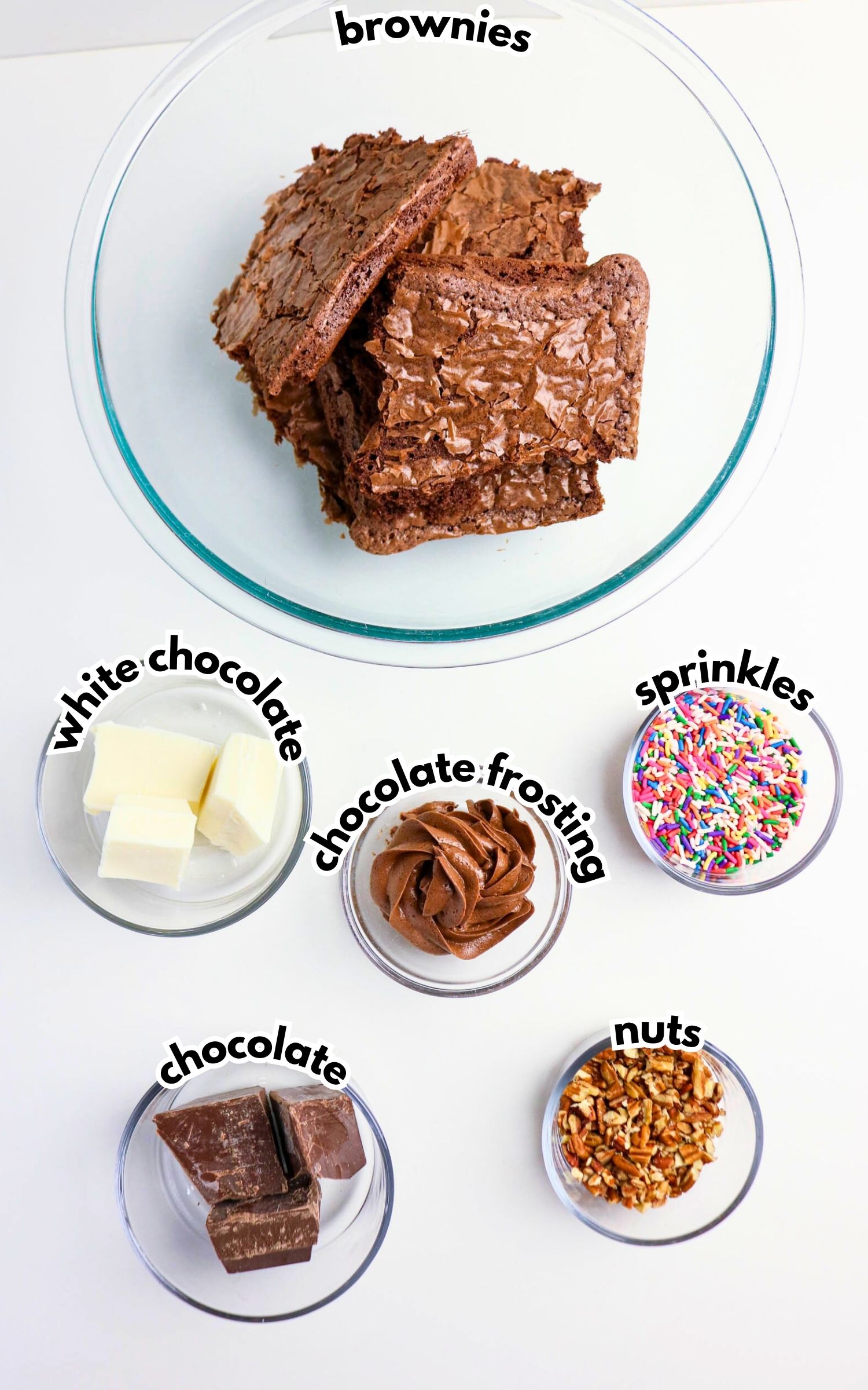 A glass bowl of brownies is surrounded by smaller bowls containing white chocolate, chocolate chunks, chocolate frosting, sprinkles, and chopped nuts.