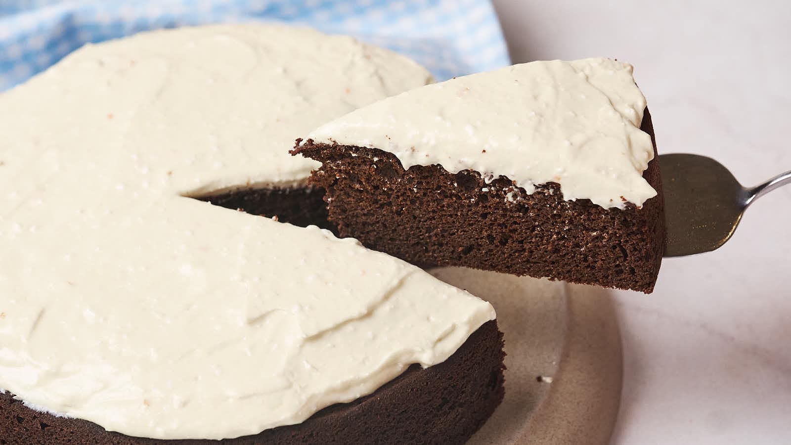 A slice of chocolate cake with white frosting is being lifted from a round cake on a plate.