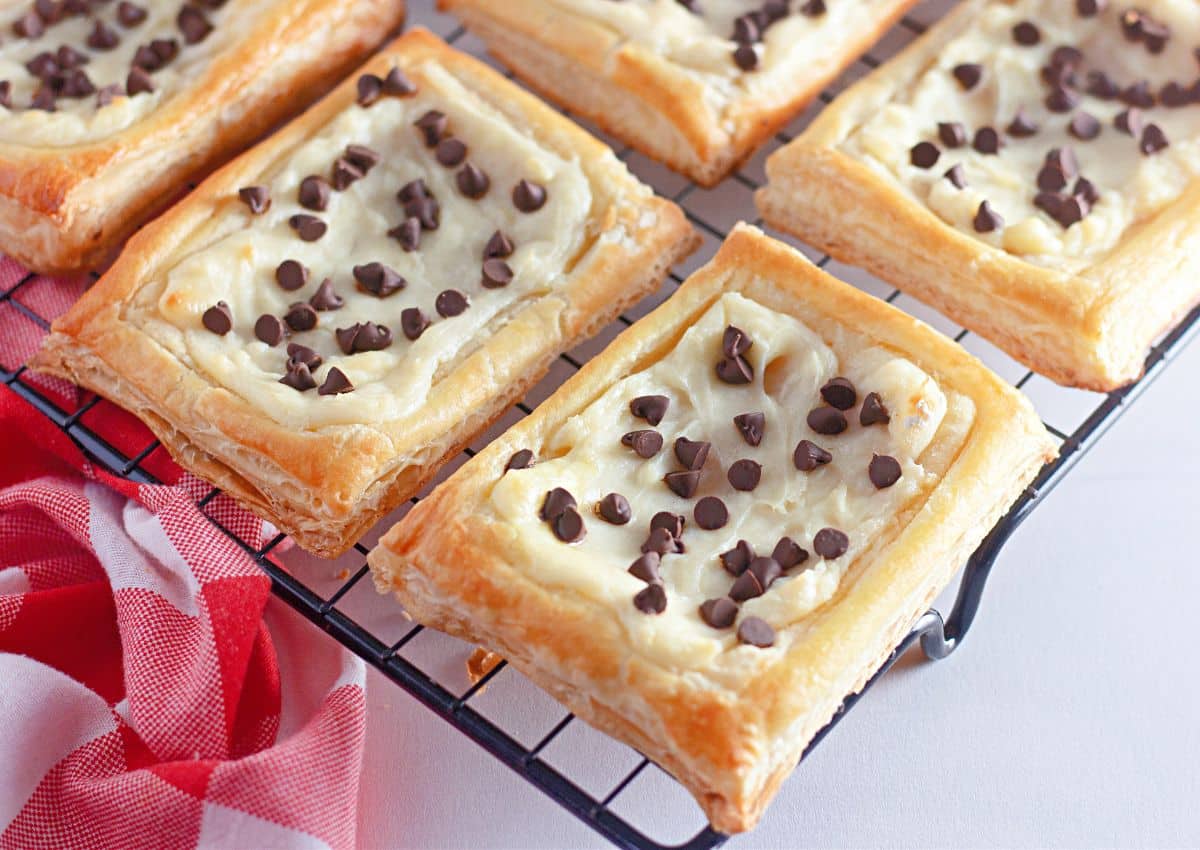 Rectangular pastries with a creamy filling and chocolate chips on top are cooling on a wire rack next to a red and white cloth.