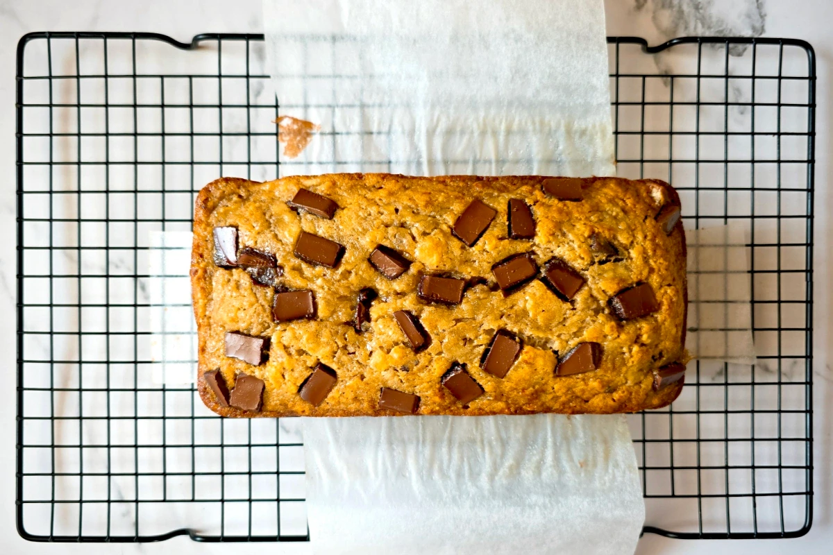 A loaf of banana bread with chocolate chunks on top sits on parchment paper over a black cooling rack.