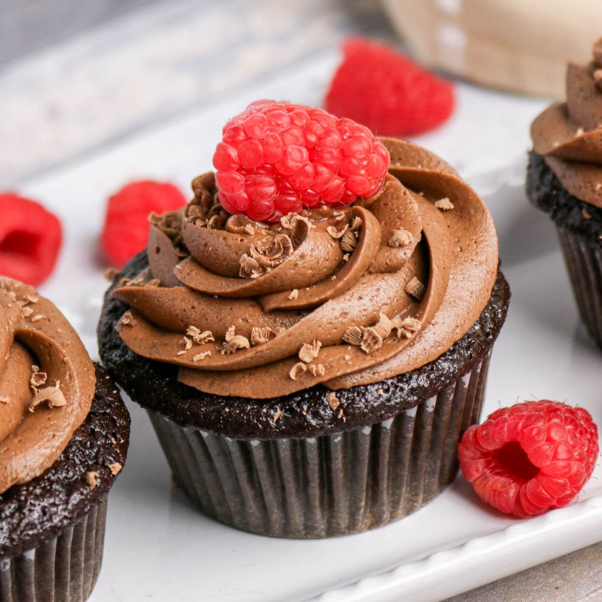 Chocolate cupcake with swirled chocolate frosting, topped with a fresh raspberry and chocolate shavings, displayed on a white plate with additional raspberries.