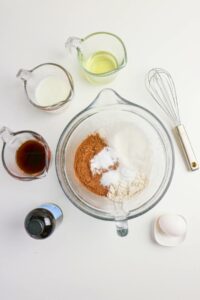 A mixing bowl with flour, cocoa powder, and baking powder, surrounded by a whisk, egg, vanilla, oil, and milk in measuring cups on a white surface.