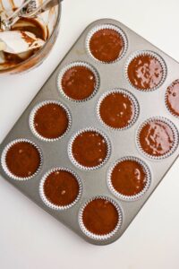A muffin tin filled with paper liners and chocolate cupcake batter, next to a bowl with remnants of batter and a spatula on a white surface.