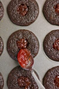 A spoon adds red jam to the center of chocolate cupcakes in a muffin tray.
