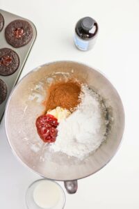 A mixing bowl with powdered sugar, cocoa powder, butter, and jam beside a muffin tin with chocolate cupcakes, a bottle of vanilla extract, and a glass of milk.