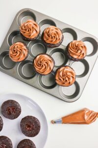 A muffin tin with chocolate cupcakes topped with swirled frosting, a plate of unfrosted cupcakes, and a piping bag filled with chocolate frosting on a white surface.