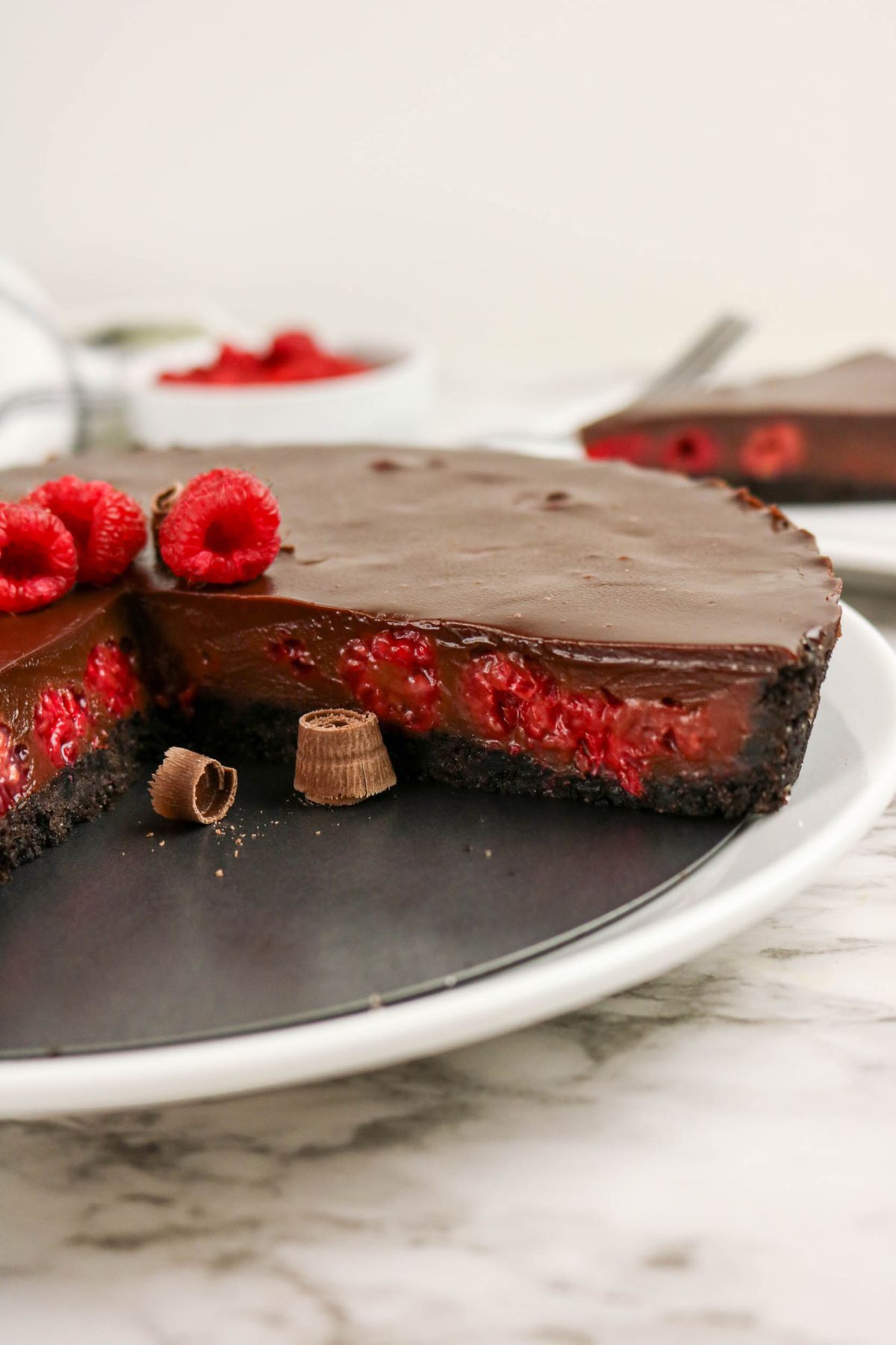 A chocolate raspberry tart with a slice removed, showing a chocolate ganache topping, raspberries inside, and a dark crust, garnished with fresh raspberries and chocolate pieces.