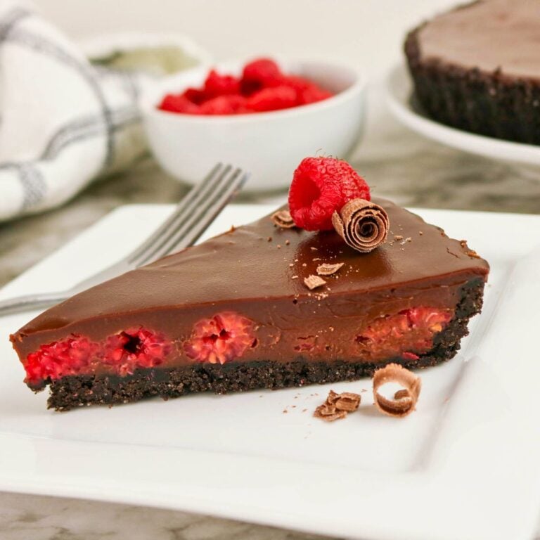 A slice of chocolate tart with raspberries and chocolate curls on top, served on a white plate with a fork.