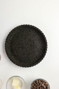 A round tart pan filled with a dark Oreo cookie crust, with bowls of butter and chocolate chips nearby on a white surface.