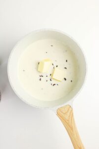 White saucepan with creamy liquid, two cubes of butter, and scattered spices on a white background.