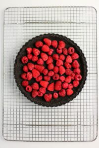 A chocolate tart topped with fresh raspberries sits on a cooling rack.