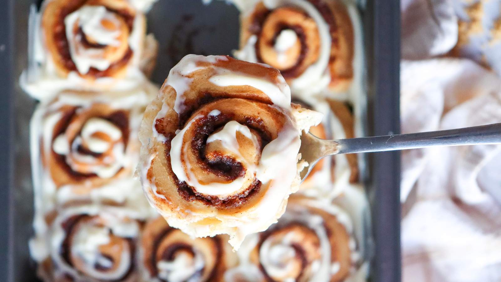 A close-up of a frosted cinnamon roll being lifted from a baking tray filled with more iced cinnamon rolls.