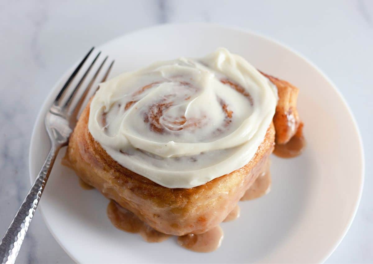 A cinnamon roll with cream cheese frosting on a white plate, with a fork beside it.