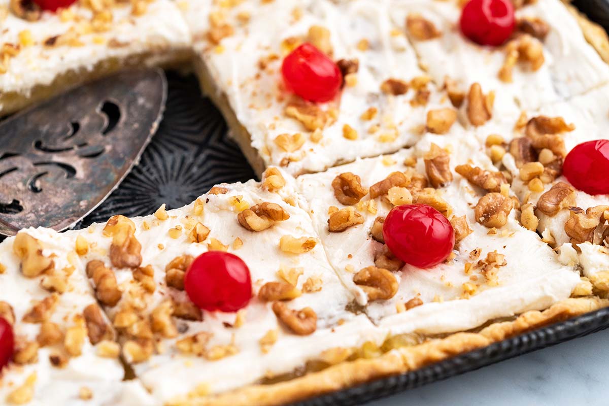 A rectangular dessert with a cream topping, chopped walnuts, and red cherries, cut into squares on a metal tray.