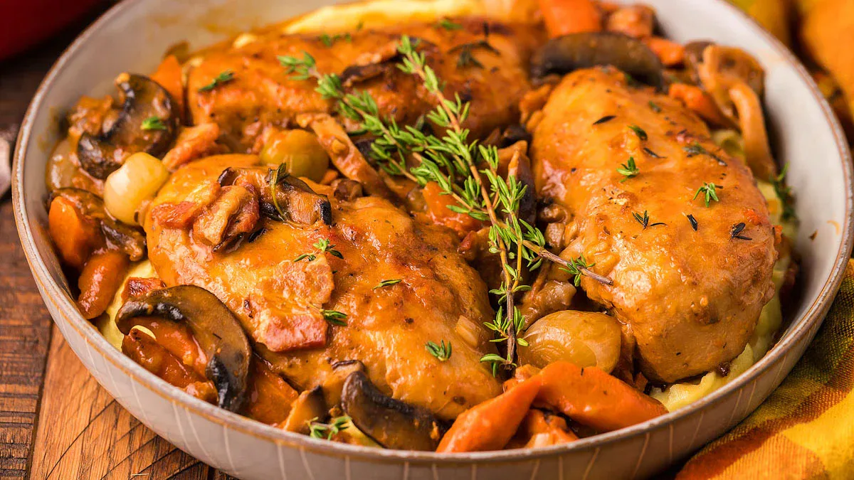 A bowl of coq au vin with braised chicken, mushrooms, carrots, onions, and fresh thyme served over mashed potatoes.