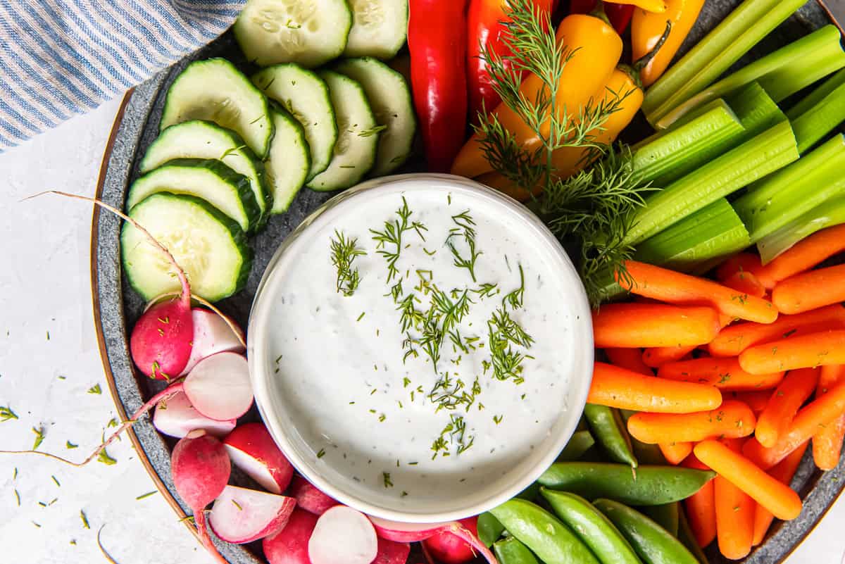 A plate of assorted fresh vegetables, including cucumber, bell peppers, celery, carrots, snap peas, and radishes, surrounds a bowl of creamy white dip garnished with dill.