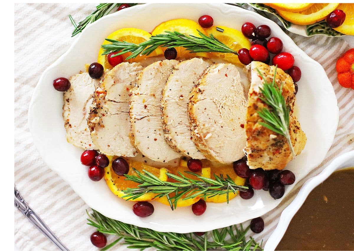Sliced roasted turkey breast garnished with orange slices, fresh rosemary, and cranberries on a white serving platter.