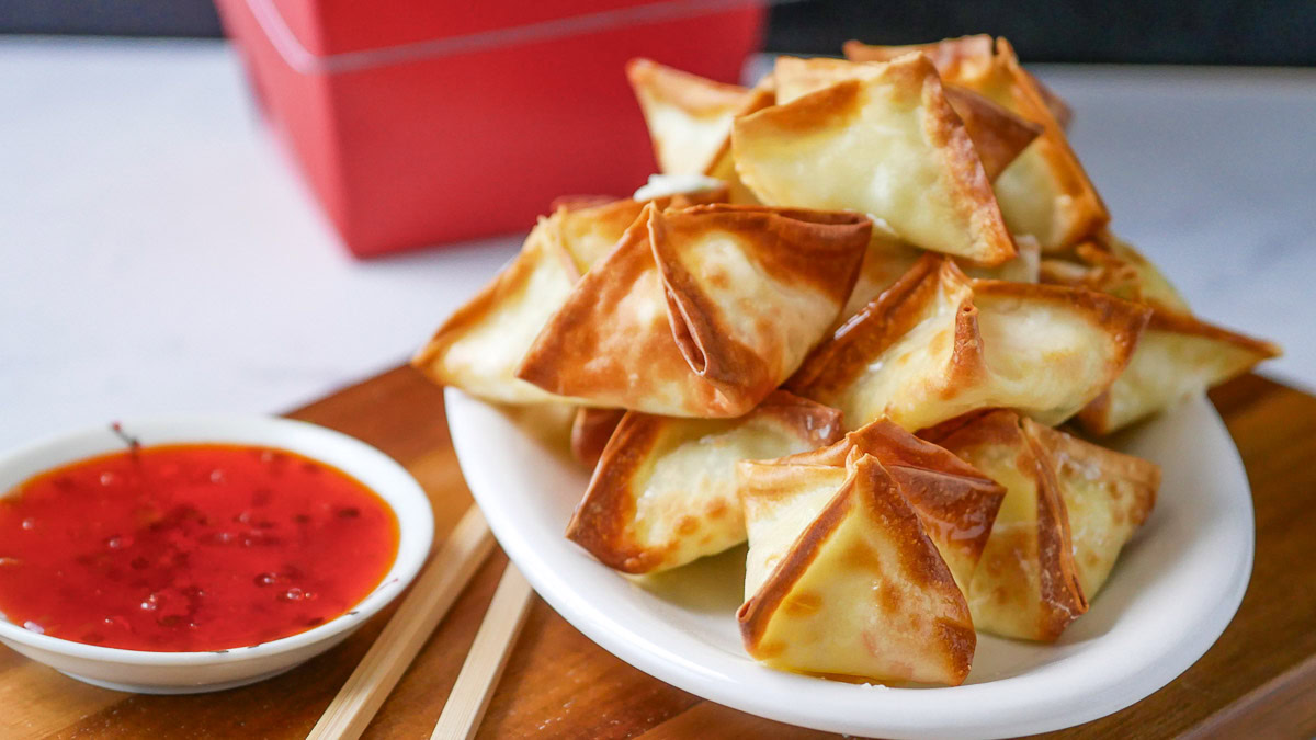A plate of crispy wontons is served with a small bowl of red dipping sauce and chopsticks, set on a wooden board.