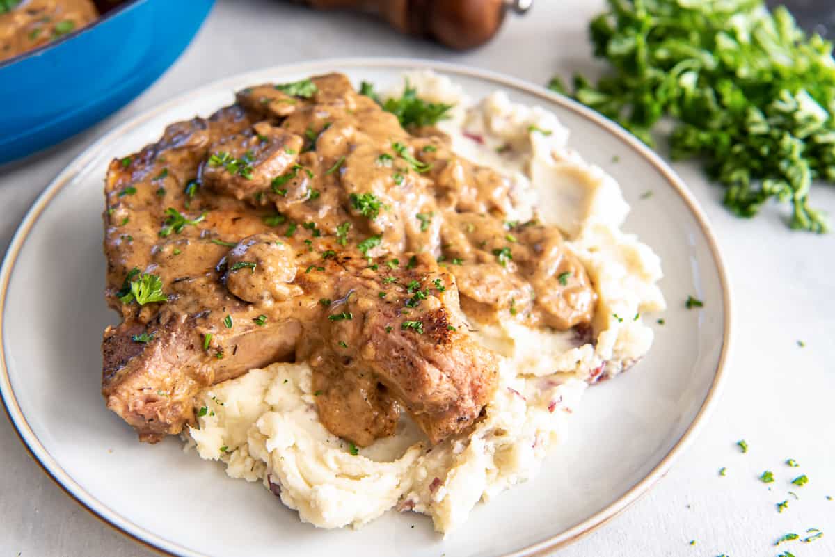 A grilled steak topped with mushroom gravy sits on a bed of mashed potatoes, garnished with chopped parsley on a white plate.