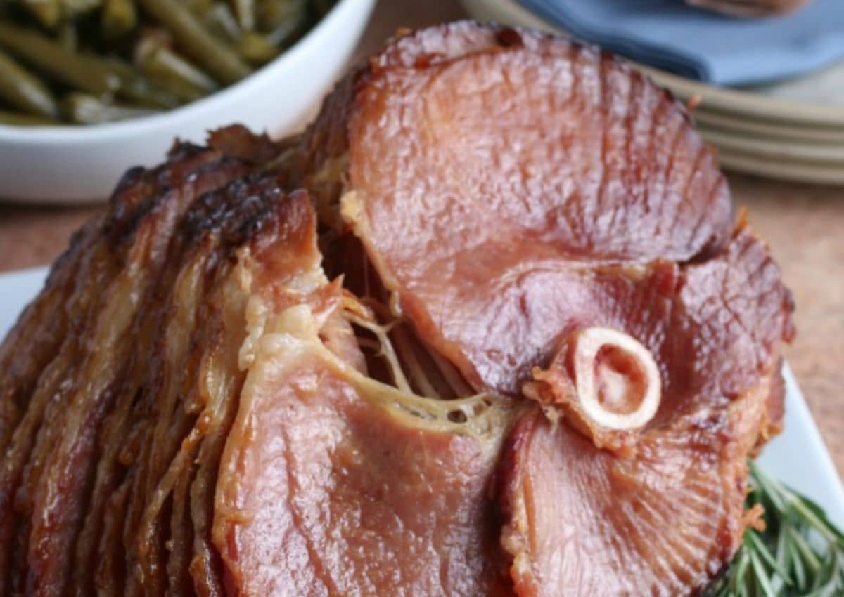 Close-up of a sliced glazed ham with a bone in the center, served on a platter with a bowl of green beans in the background.